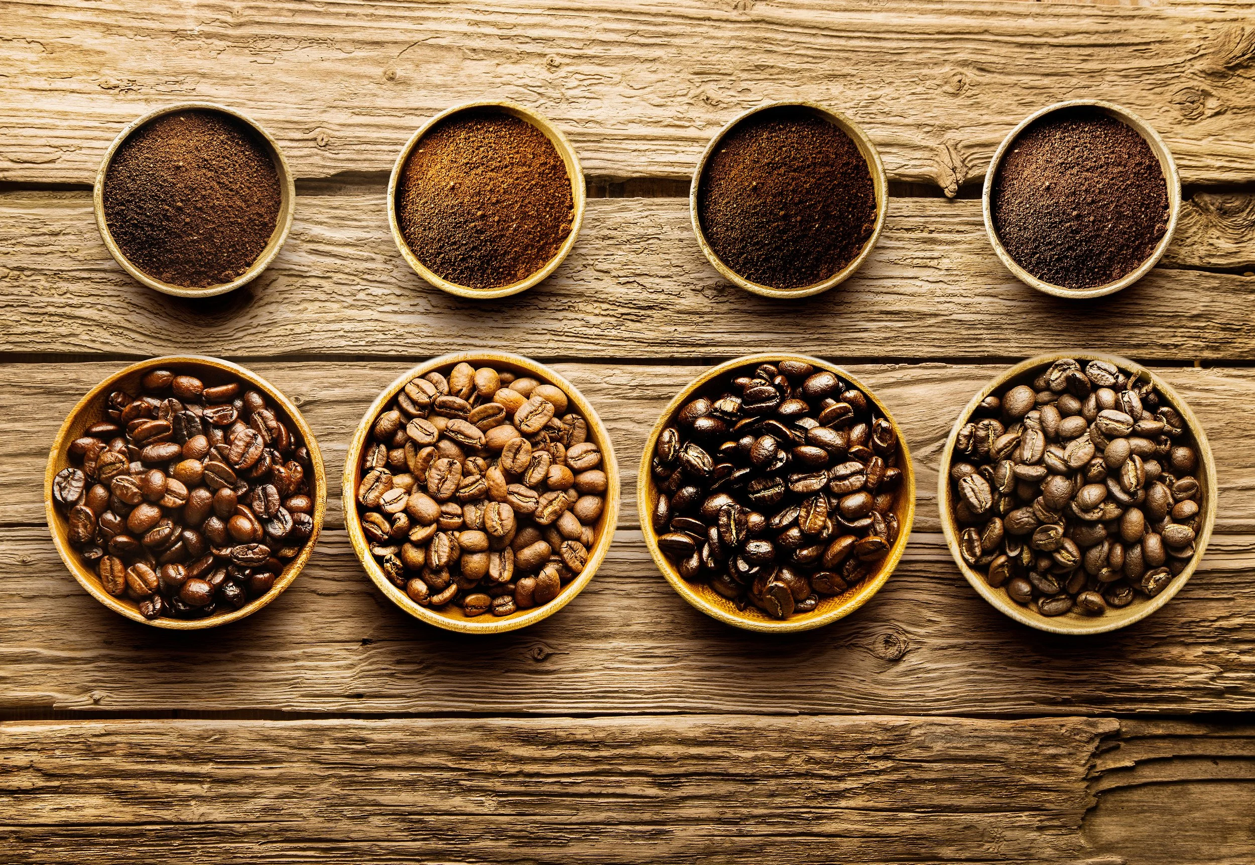 Four wooden bowls of green and roasted coffee beans alongside grounds in different roast levels, on a wooden bench
